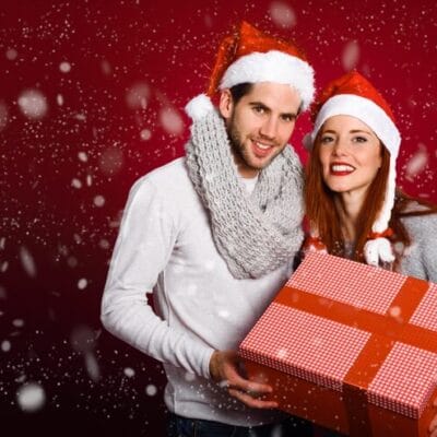 couple-with-christmas-gift-dressed-with-winter-clothes-santa-hat-snow-red-background_23-2147576866