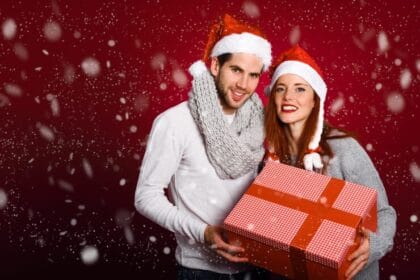 couple-with-christmas-gift-dressed-with-winter-clothes-santa-hat-snow-red-background_23-2147576866