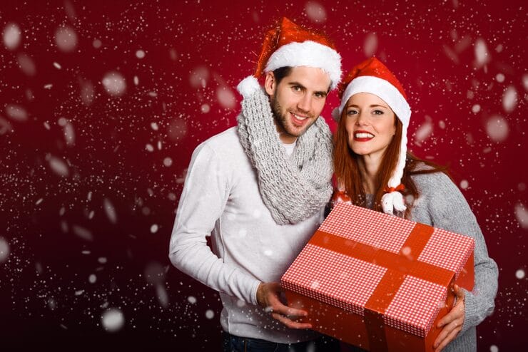 couple-with-christmas-gift-dressed-with-winter-clothes-santa-hat-snow-red-background_23-2147576866